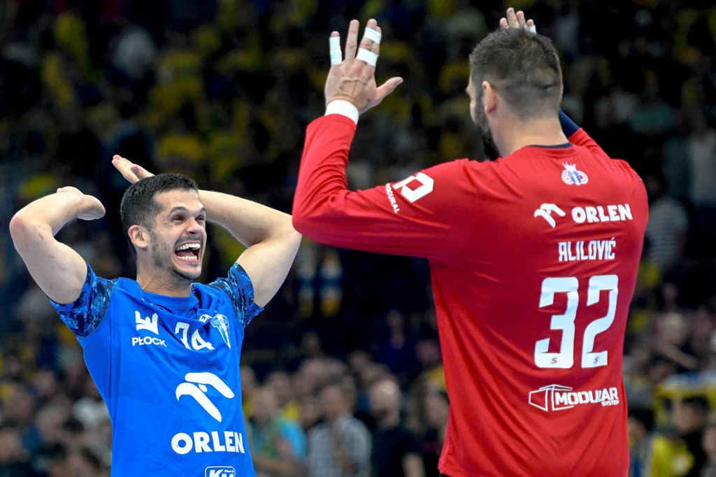 Dwóch zawodników drużyny piłki ręcznej ORLEN świętuje sukces na boisku, jeden ubrany w niebieską koszulkę, drugi w czerwony trykot z napisem ORLEN i numerem 32, wyrażają radość unosząc ręce do góry.