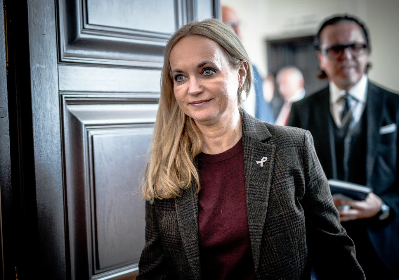 Kobieta w jasnych włosach, ubrana w ciemną marynarkę w kratę i bordową bluzkę, stoi na pierwszym planie w pomieszczeniu. Na jej marynarce widoczna jest przypinka w kształcie wstążki. W tle widoczny jest mężczyzna w garniturze oraz rozmyte postaci innyc...