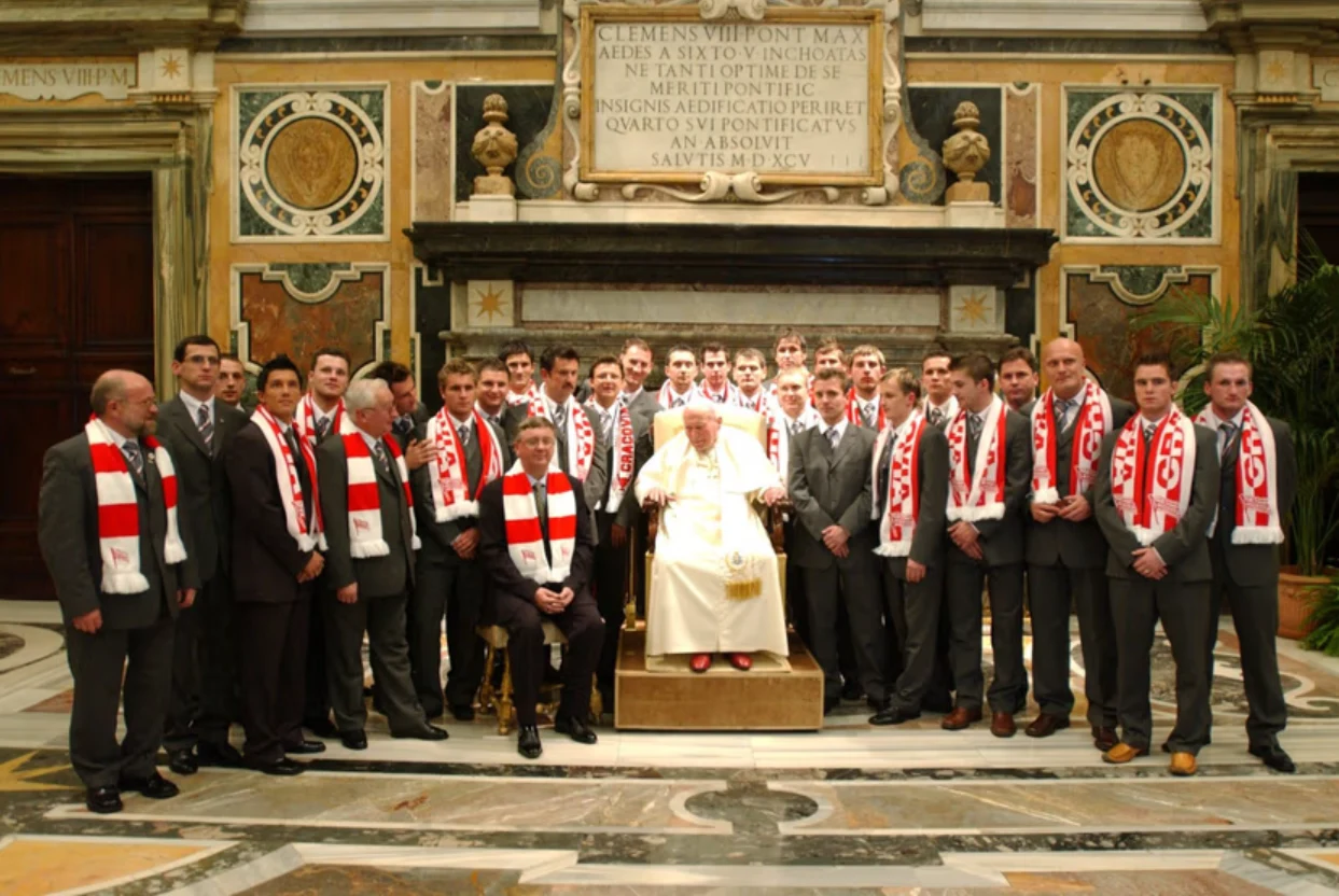 Jan Paweł II z drużyną Cracovii, 04.01.2005 r. Grupa mężczyzn ubranych w garnitury i czerwono-białe szaliki stoi wokół starszego mężczyzny w białych szatach siedzącego na tronie w bogato zdobionym wnętrzu pałacowym.