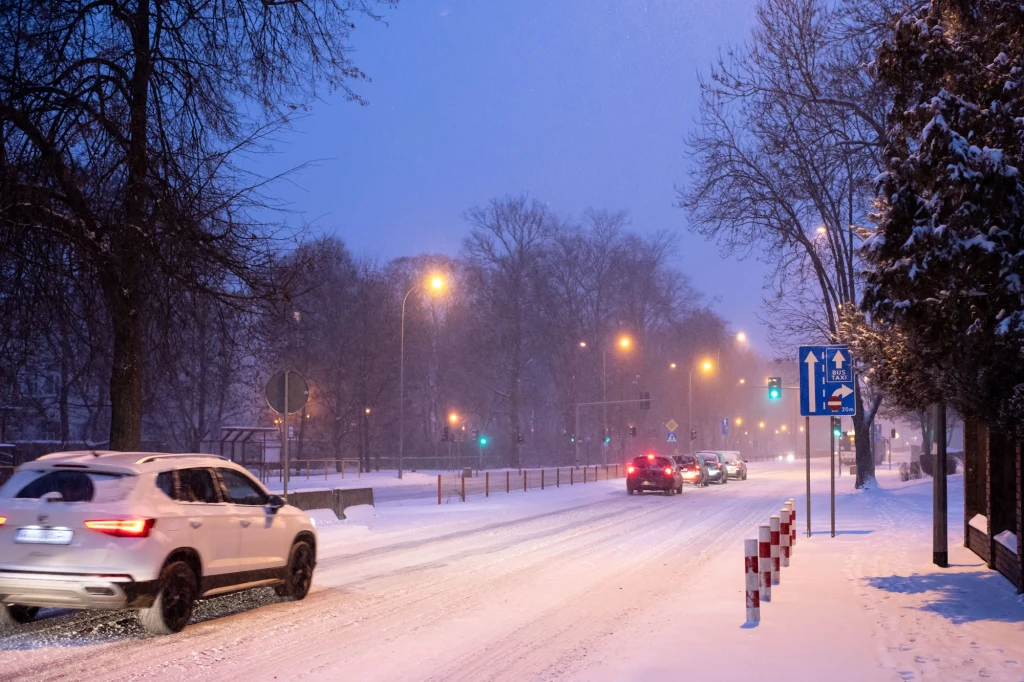 Zaśnieżona ulica w mieście o zmierzchu, sznur samochodów poruszających się w obu kierunkach, światła latarni i sygnalizacji świetlnej rozświetlają zimowy pejzaż, po prawej stronie chodnik pokryty śniegiem i znaki drogowe.