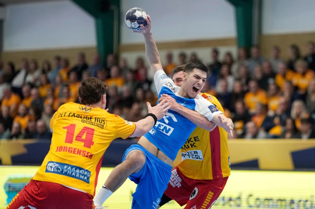 Zawodnik w niebieskim stroju wykonuje dynamiczny rzut piłką ręczną, będąc otoczonym przez dwóch obrońców w żółto-czerwonych strojach. W tle widoczna publiczność uważnie obserwuje przebieg meczu.