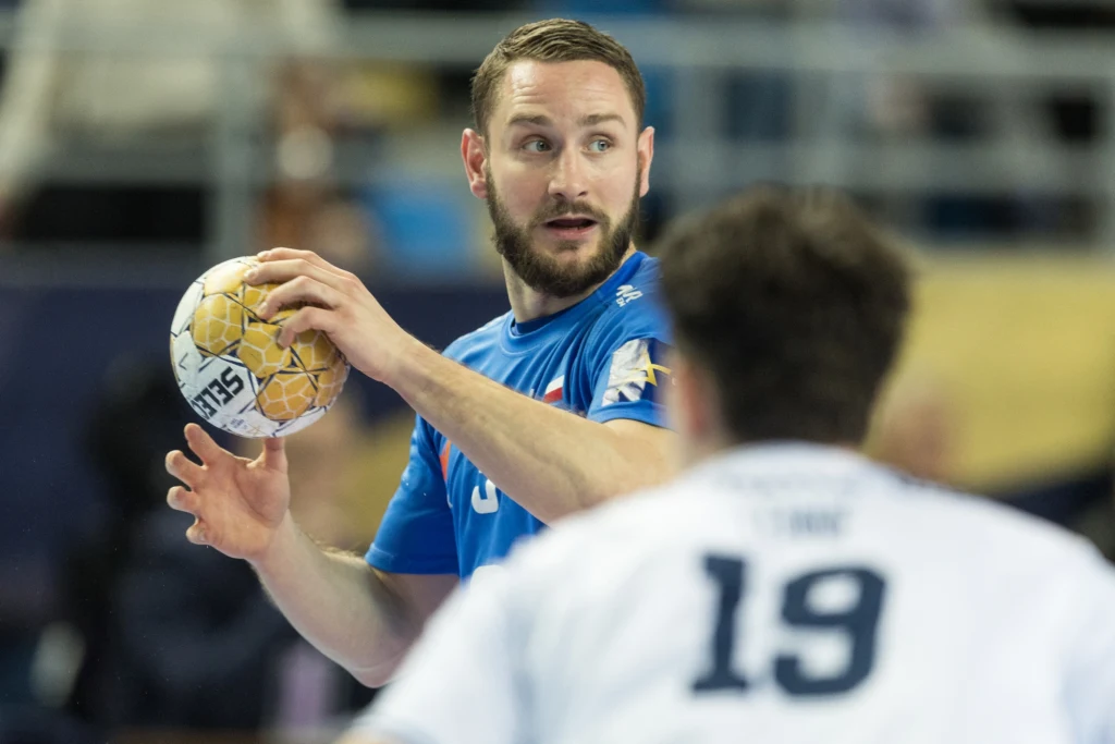 Zawodnik w niebieskiej koszulce z piłką w ręku, koncentrujący się na grze podczas meczu piłki ręcznej, w tle widać nieostrego przeciwnika w białym stroju oznaczonego numerem 19.