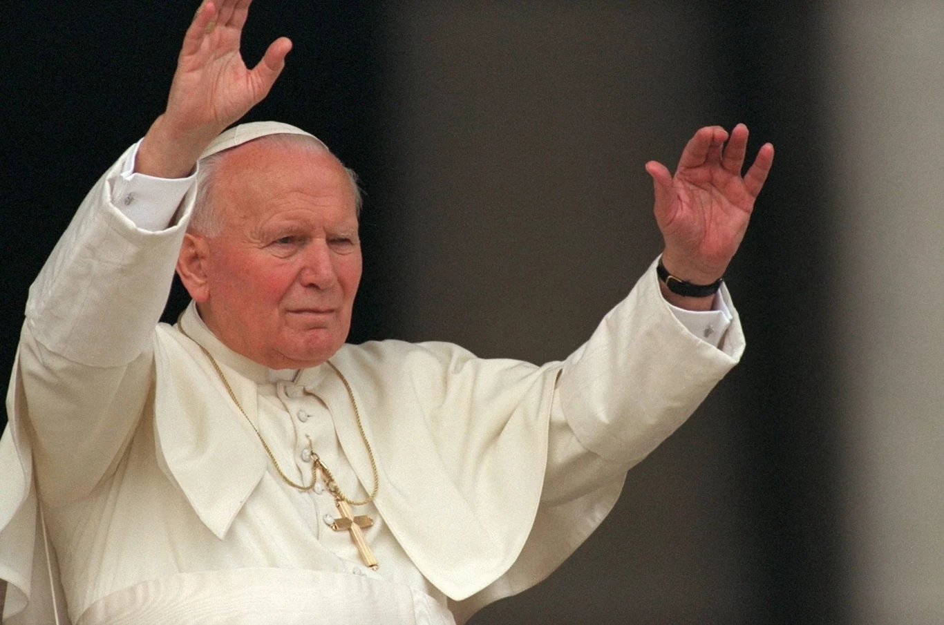 Jan Paweł II, 02.10.1996 r. Starszy mężczyzna w białej szacie papieskiej i białej piusce podnosi obie ręce w geście powitania, na szyi zawieszony duży złoty krzyż, tło ciemne i neutralne.