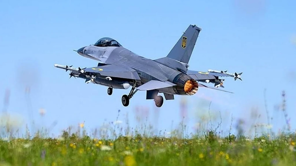 Myśliwiec wojskowy F-16 w locie tuż nad łąką, z widocznym pracującym silnikiem odrzutowym i uzbrojeniem pod skrzydłami na tle błękitnego nieba.