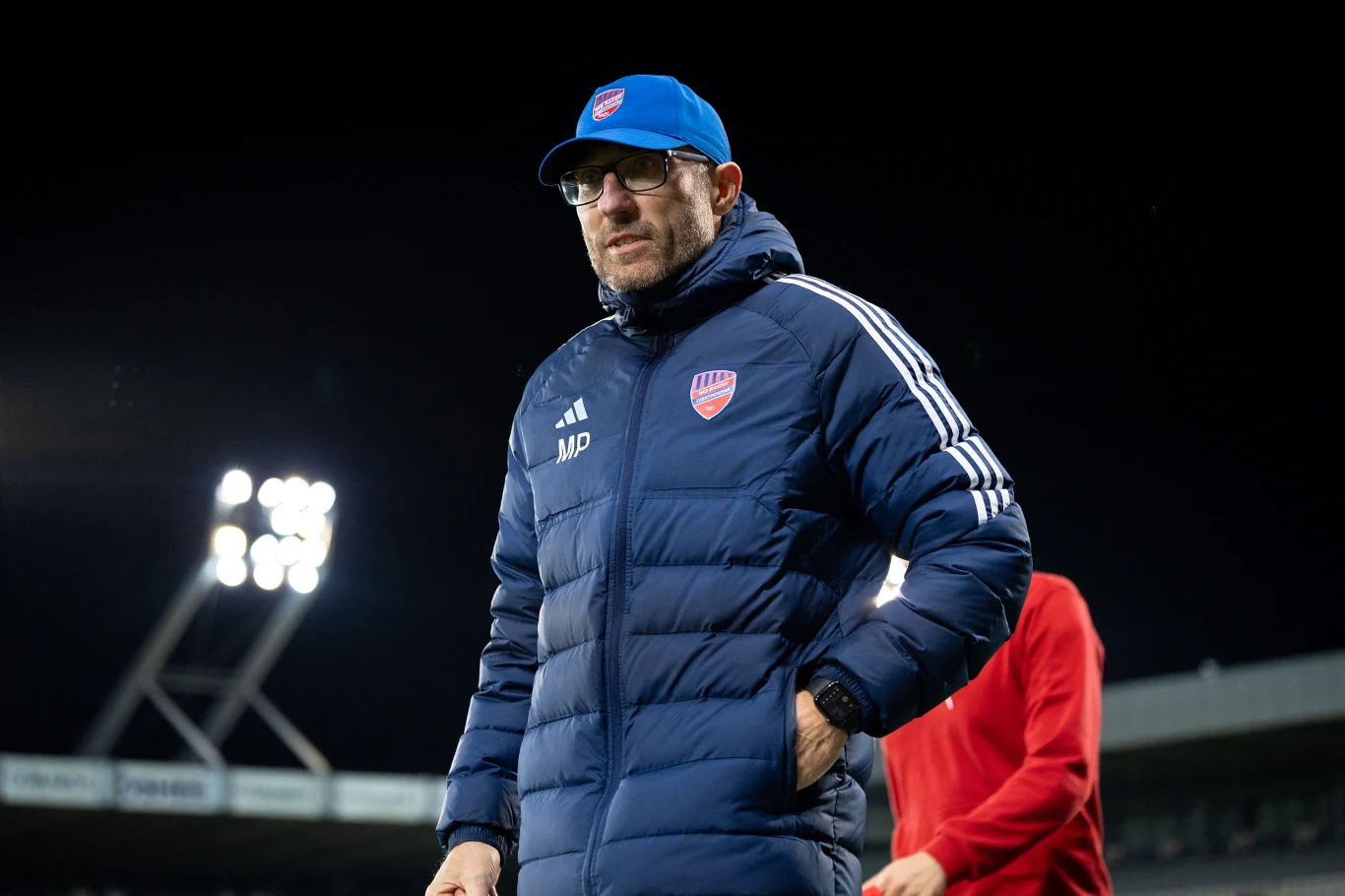 Marek Papszun Mężczyzna ubrany w sportową kurtkę i czapkę z daszkiem, z logo klubu na piersi, stoi na tle oświetlonego stadionu sportowego. W tle widoczne jasne reflektory stadionowe oraz postać ubrana na czerwono.