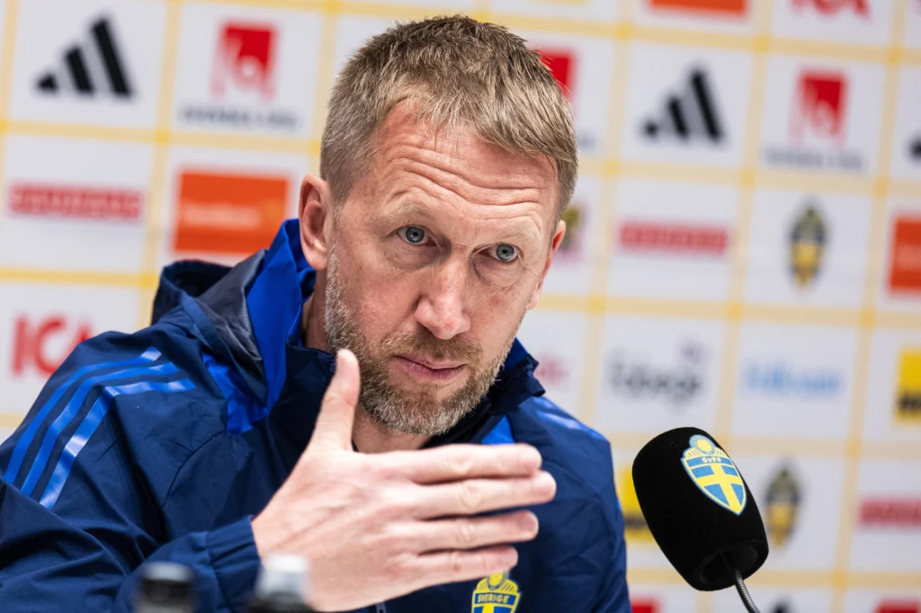 Graham Potter, selekcjoner reprezentacji Szwecji Mężczyzna w sportowej kurtce z logo Szwecji gestykuluje podczas konferencji prasowej, przed nim mikrofon z emblematem szwedzkiej federacji piłkarskiej, w tle ścianka z logo sponsorów.