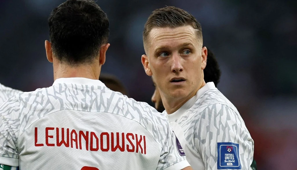 Do triumfu w fazie barażowej poprowadzić nas mają liderzy kadry - Robert Lewandowski i Piotr Zieliński Dwóch piłkarzy w białych koszulkach reprezentacji Polski na boisku, jeden z nich odwrócony tyłem z widocznym nazwiskiem Lewandowski, drugi spogląda w bok, skupiony wyraz twarzy.