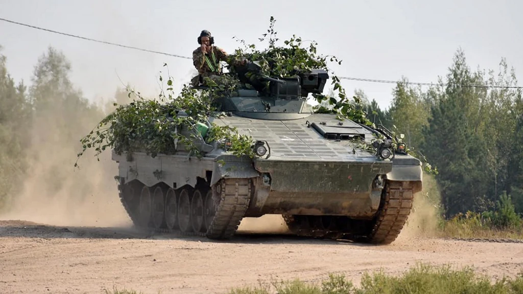 Opancerzony pojazd wojskowy jadący po piaszczystej drodze, częściowo zamaskowany gałęziami, otoczony przez roślinność leśną, żołnierz widoczny w górnej części pojazdu.