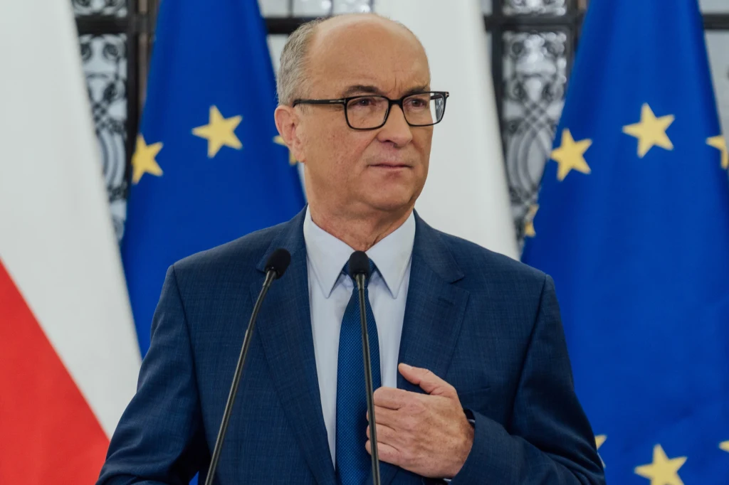 Mężczyzna w garniturze i okularach stoi przy mównicy, w tle widoczne są flagi Unii Europejskiej oraz Polski.