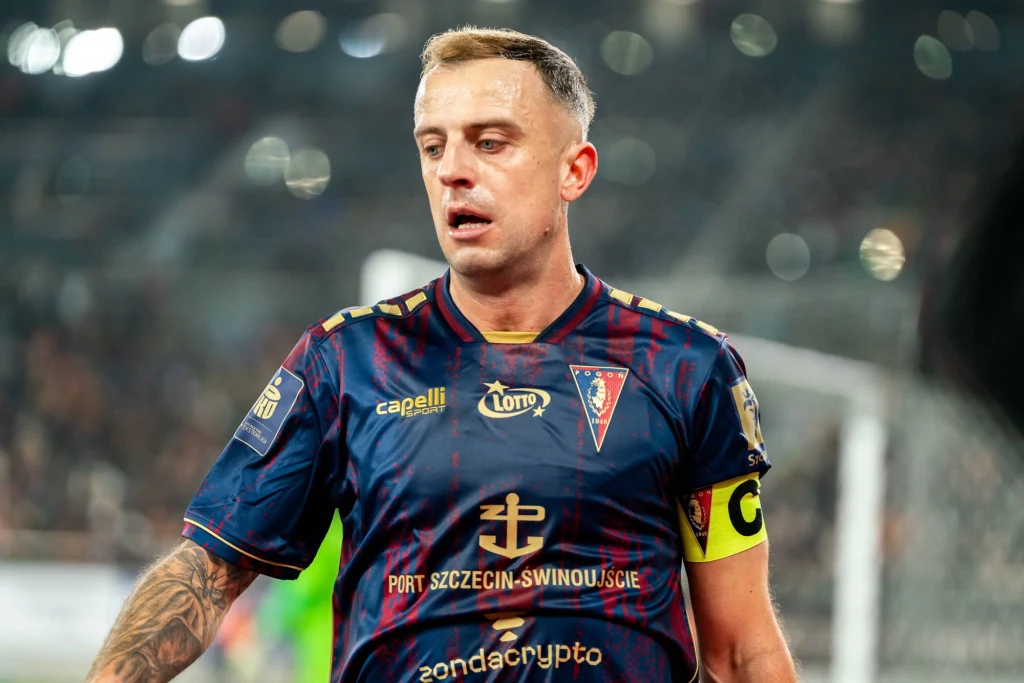 Kamil Grosicki Piłkarz w granatowo-bordowej koszulce z emblematem klubu Pogoń Szczecin, opaska kapitana na ramieniu, widok podczas meczu piłkarskiego, rozmyte tło stadionu.