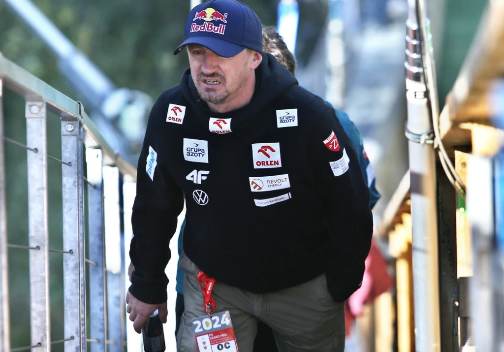 Mężczyzna w czapce z daszkiem i bluzie sportowej z licznymi logotypami sponsorów wspina się po schodach na zewnątrz, trzymając w ręku butelkę. Do bluzy przypięta jest akredytacja z roku 2024.