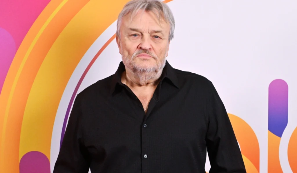 Starszy mężczyzna w czarnej koszuli, stojący na tle kolorowej grafiki z okręgami i liniami w odcieniach pomarańczu, różu i fioletu.