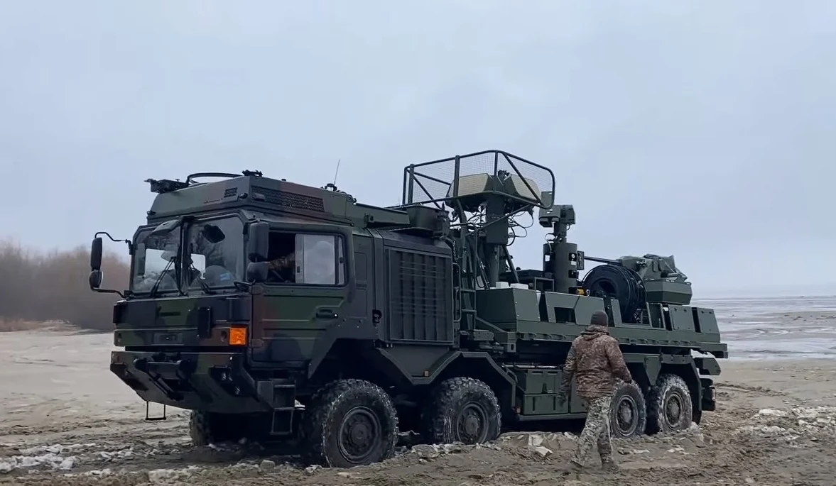 Nowa warstwa obrony przeciwdronowej Ukrainy. Brytyjski Terrahawk Paladin już na froncie Wojskowy pojazd opancerzony wyposażony w radar i sprzęt komunikacyjny, zatrzymany na błotnistym terenie, obok znajduje się żołnierz w mundurze polowym.