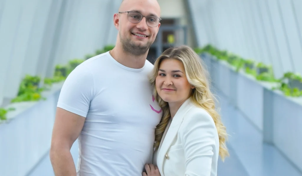 Mężczyzna w białej koszulce i kobieta w białej marynarce stoją razem na jasnym przejściu z przeszklonymi ścianami oraz zielonymi ozdobnymi roślinami po bokach. Oboje uśmiechnięci patrzą w kierunku aparatu.