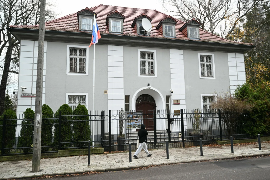 Szara kamienica z czerwonym dachem, rosyjską flagą na maszcie i metalowym ogrodzeniem. Przed budynkiem przechodzi pojedyncza osoba, w tle widoczne drzewa bez liści.