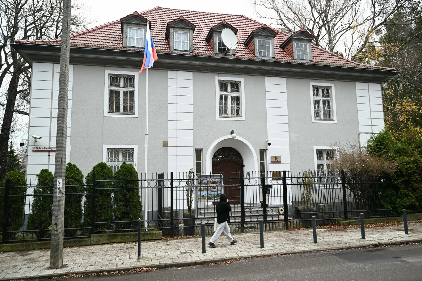Konsulat Rosji w Gdański z ogromnym zadłużeniem Szara kamienica z czerwonym dachem, rosyjską flagą na maszcie i metalowym ogrodzeniem. Przed budynkiem przechodzi pojedyncza osoba, w tle widoczne drzewa bez liści.