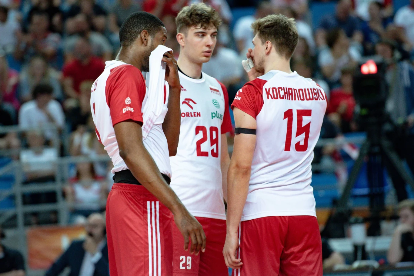 Trzech siatkarzy drużyny narodowej Polski w trakcie rozmowy na boisku, ubranych w biało-czerwone stroje meczowe, otoczeni przez kibiców na trybunach.