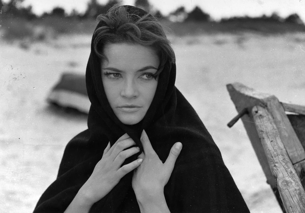 Młoda kobieta z ciemnym szalem otulającym głowę i ramiona spogląda zamyślonym wzrokiem na bok, stojąc na tle rozmytego krajobrazu z łodzią i drzewami w oddali.