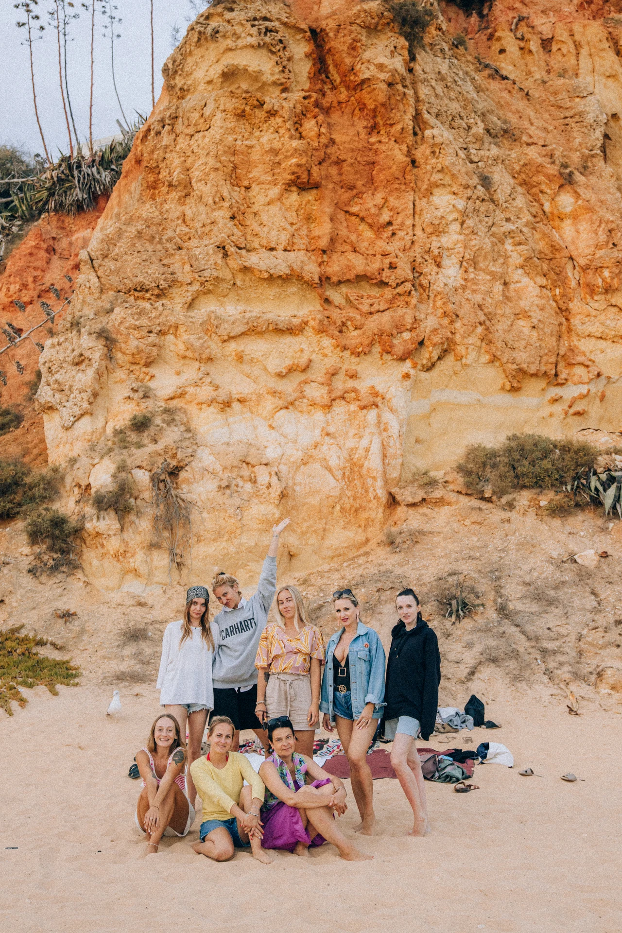 Polka organizuje “babskie wyjazdy” do Portugalii Grupa kobiet pozuje razem na piaszczystej plaży przed wysoką, pomarańczowo-żółtą skałą. Kilka osób stoi, inne siedzą, wszyscy wyglądają na zrelaksowane i uśmiechnięte. W tle można dostrzec roślinność rosnącą na skale i ubrania rozłożone na piasku.