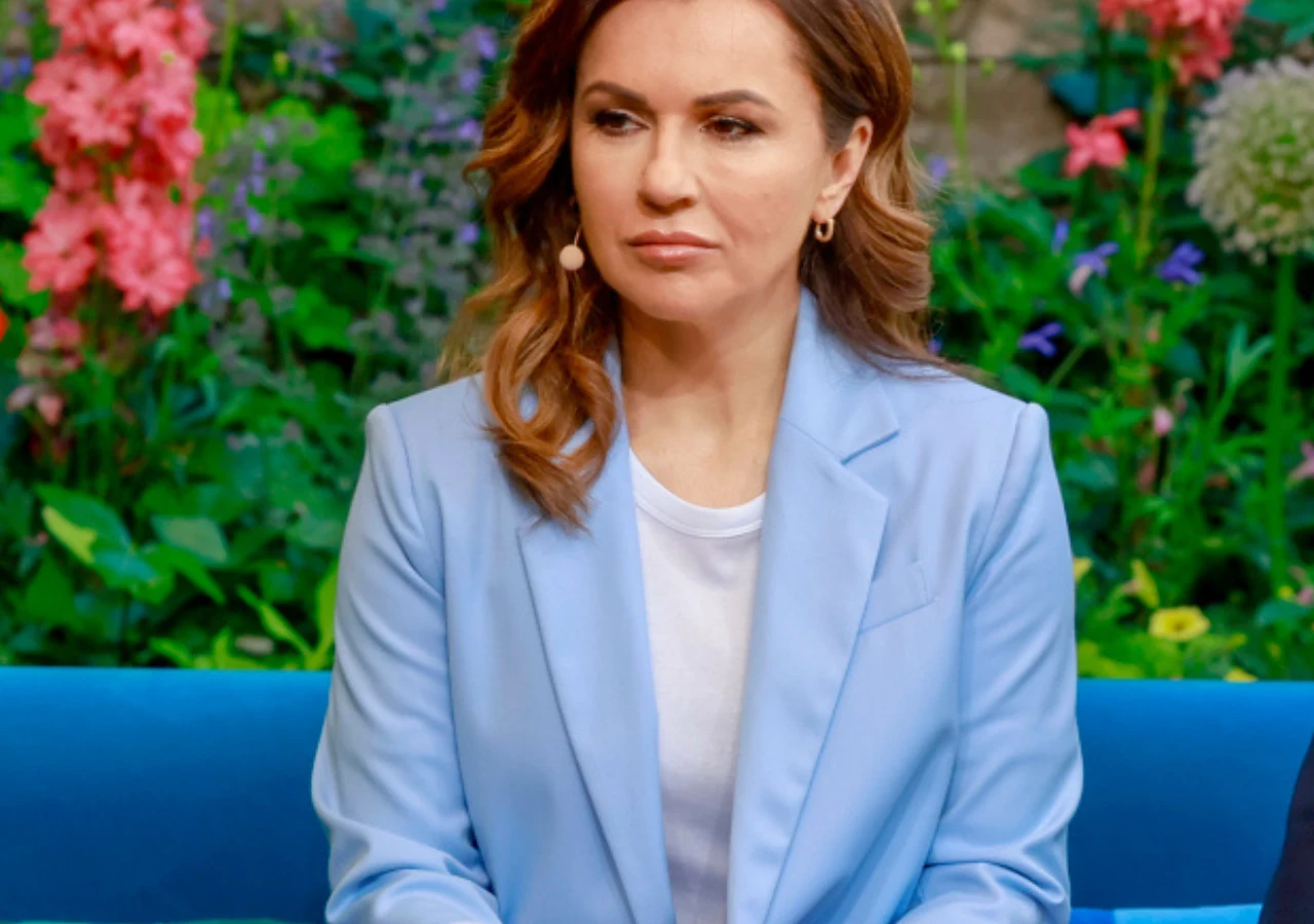 Beata Tadla Kobieta o brązowych włosach ubrana w jasnoniebieską marynarkę i białą koszulkę siedzi na niebieskim tle, w tle widoczne kolorowe kwiaty ogrodowe.