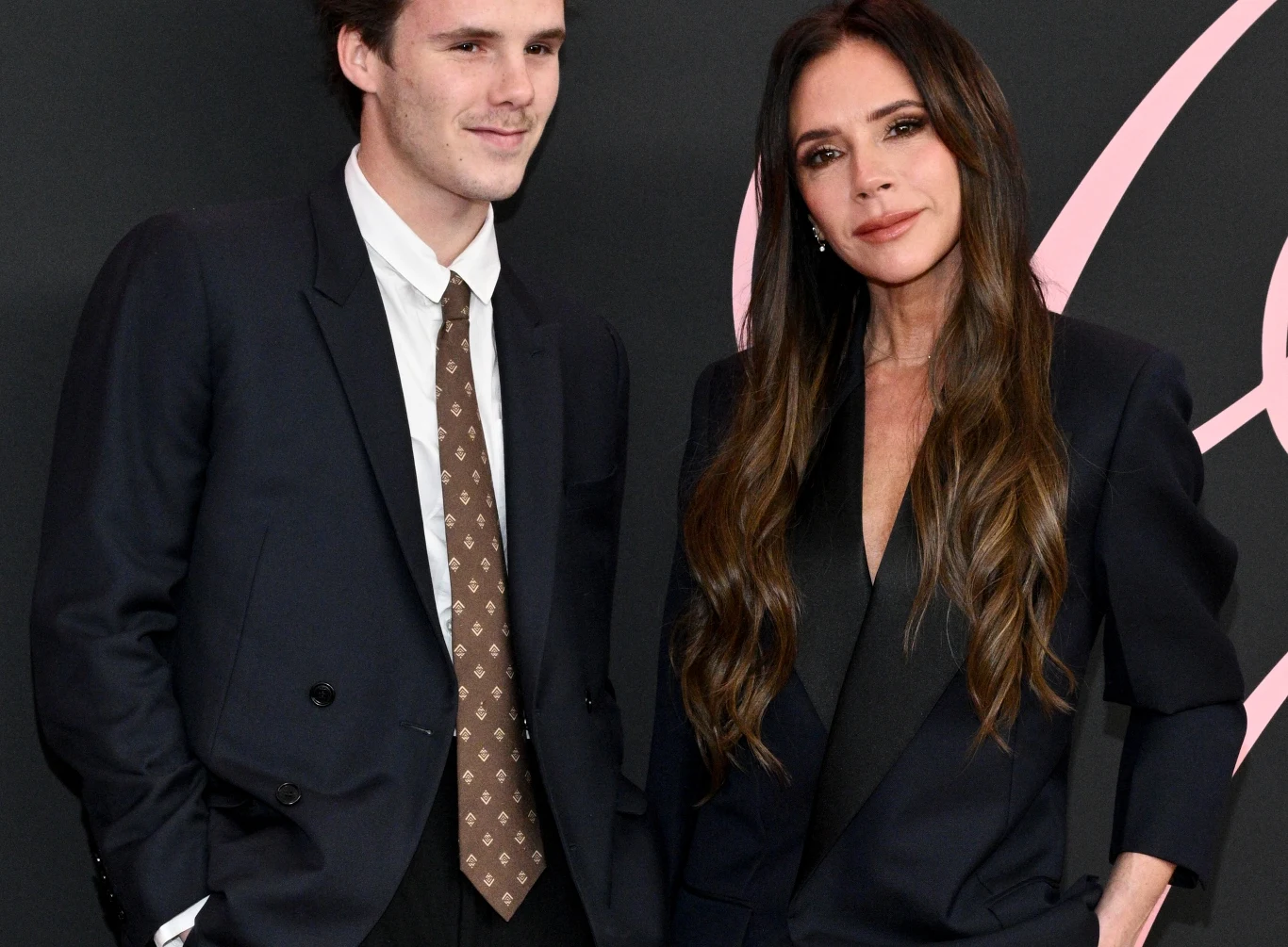 Victoria Beckham z synem zaśpiewała kultowy utwór Spice Girls Mężczyzna i kobieta ubrani elegancko, stoją razem na tle ciemnej ściany z dekoracyjnym wzorem.