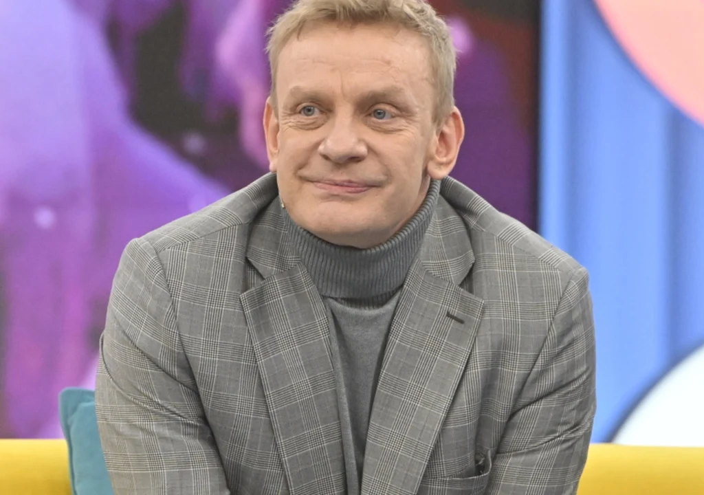 Mężczyzna w szarej marynarce w kratę i golfie siedzi na żółtej sofie, w tle kolorowe, rozmyte elementy dekoracji studyjnej.