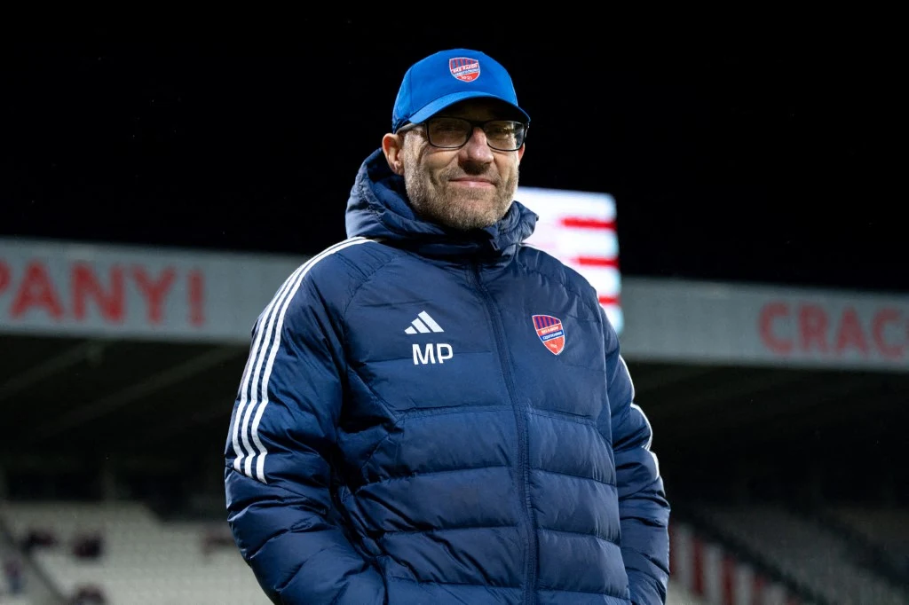 Marek Papszun został trenerem Legii Warszawa Mężczyzna w sportowej kurtce i czapce z daszkiem, ubrany w zimowe ubranie klubowe z herbem piłkarskim oraz inicjałami MP, stoi na stadionie piłkarskim, w tle trybuny i częściowo widoczny napis reklamowy.