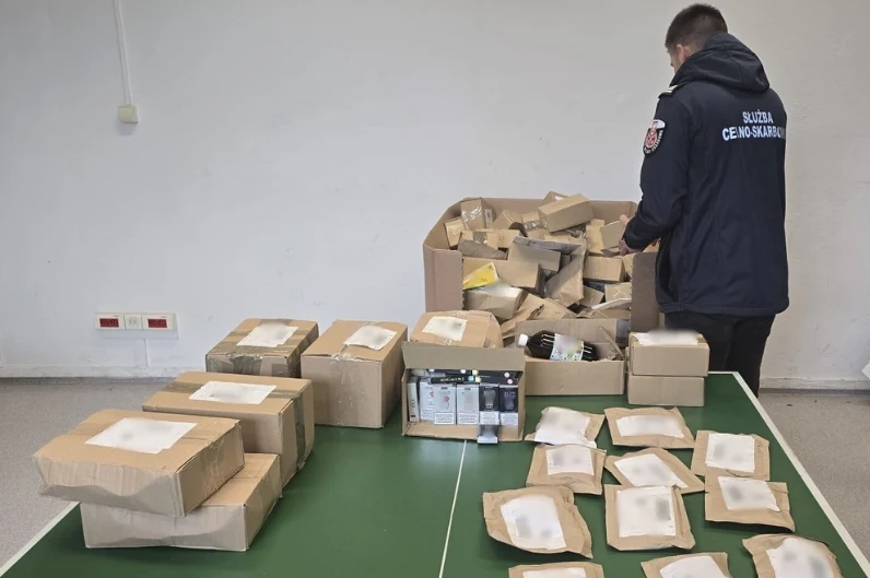Funkcjonariusz służby celno-skarbowej przeszukuje i segreguje dużą ilość kartonowych paczek rozłożonych na stole oraz w tle, w pudełkach znajdują się różne przedmioty i opakowania.