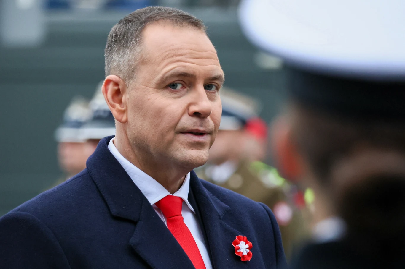 Prezydent RP Karol Nawrocki Mężczyzna w eleganckim płaszczu i czerwonym krawacie, z przypiętym biało-czerwonym znakiem rozpoznawczym, stoi na tle rozmytych sylwetek wojskowych w mundurach i innych uczestników uroczystości.
