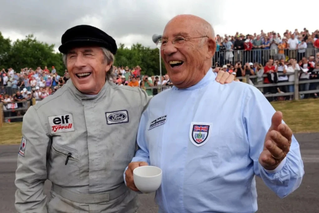 Dwóch starszych mężczyzn uśmiecha się i rozmawia ze sobą na tle dużej grupy ludzi. Jeden z nich ubrany jest w wyścigowy kombinezon i czapkę, drugi ma jasną koszulę z emblematem i trzyma filiżankę. Pogodna atmosfera i tłum zgromadzony za barierką podkre...
