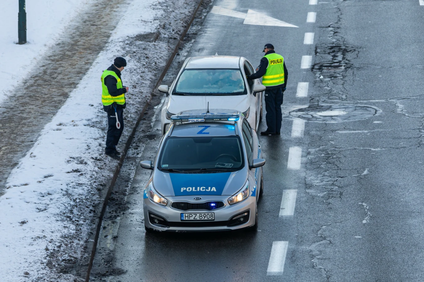 Za jazdę z zaparowanymi szybami grozi mandat. Warto o tym pamiętać Dwóch policjantów w żółtych kamizelkach odblaskowych dokonuje kontroli drogowej białego samochodu na zimowej, zaśnieżonej ulicy, przed pojazdem stoi radiowóz policyjny.