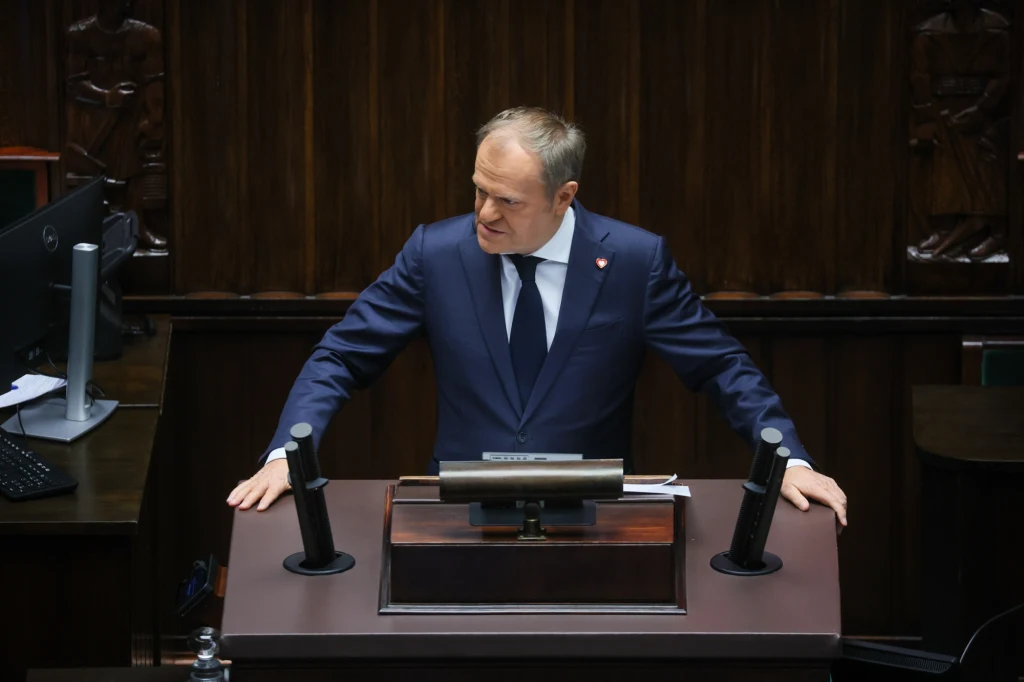 Donald Tusk Mężczyzna w garniturze stoi za mównicą w sali obrad, opierając ręce o pulpit i zwracając się do zgromadzonych. W tle widoczne są ciemne, drewniane elementy wystroju oraz sprzęt elektroniczny po lewej stronie.