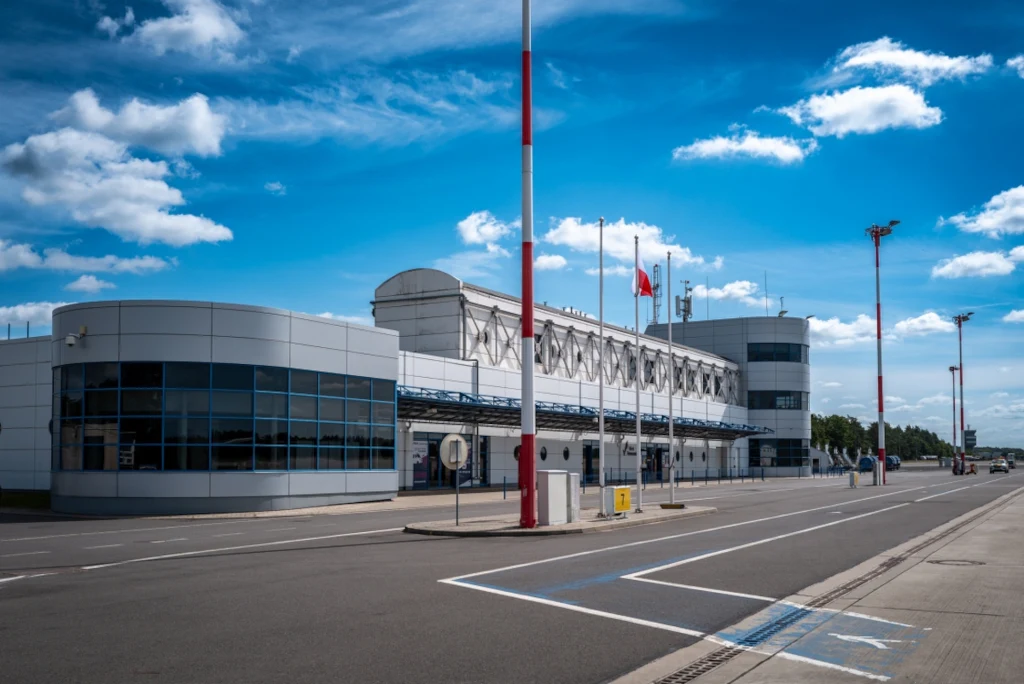 Lotnisko Szczecin-Goleniów stawia tymczasowy terminal pasażerski. Budynek portu lotniczego o nowoczesnej architekturze z przeszklonymi ścianami i polską flagą na jednym z masztów, szeroka jezdnia przed terminalem, niebo ze sporą ilością białych chmur.