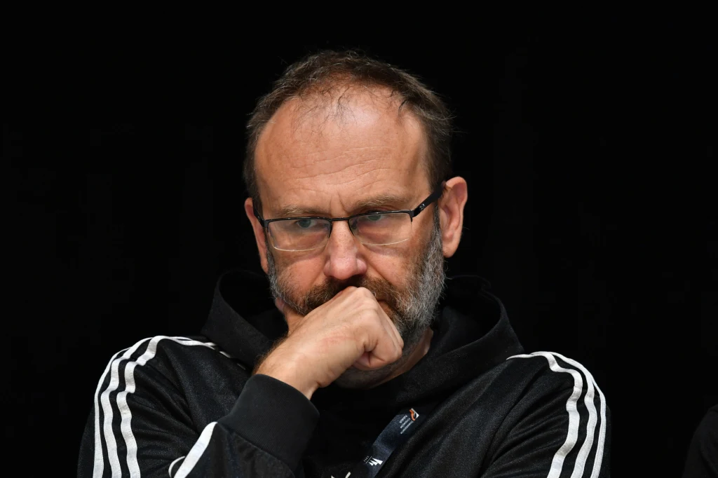 Mężczyzna w średnim wieku z brodą i okularami, siedzący ze skrzyżowanymi ramionami i opartą na ręku głową, ubrany w ciemną bluzę z białymi paskami, skupiony i zamyślony na ciemnym tle.