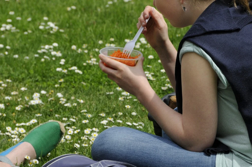 Osoba siedzi na trawie w otoczeniu kwiatów stokrotek, trzymając plastikową miseczkę z jedzeniem i jedząc widelcem. Ubrana w jasnoniebieskie spodnie i zielone buty, z widoczną częścią ramienia oraz plecak obok.