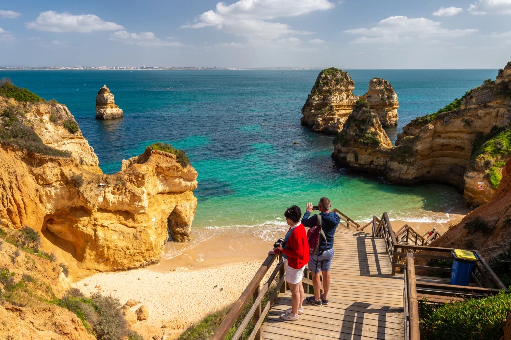 Krajobrazy Algarve na długo pozostają w pamięci Drewniane schody prowadzące na piaszczystą plażę ze skalistymi klifami, turkusowa woda oceanu, dwoje ludzi podziwiających widok oraz charakterystyczne formacje skalne wystające z morza.