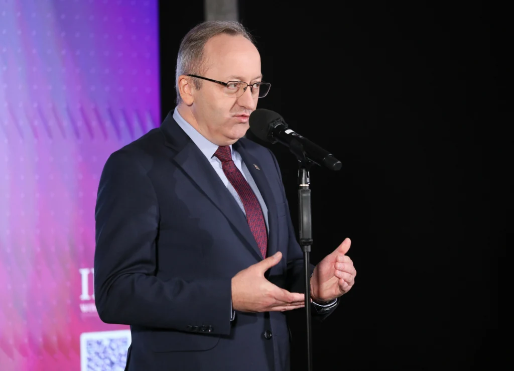 Karol Polejowski Mężczyzna w garniturze i okularach przemawia do mikrofonu na tle gradientowej planszy z elementami graficznymi, gestykulując podczas wypowiedzi.