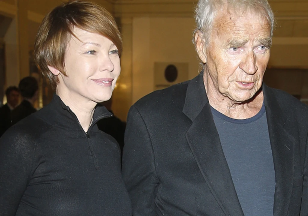 Dwie osoby w średnim i starszym wieku, kobieta i mężczyzna, stojące obok siebie w eleganckich, ciemnych ubraniach, na tle jasnego wnętrza.