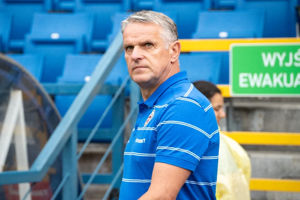 Mężczyzna w niebieskiej koszulce polo z białymi paskami stoi na tle stadionowych niebieskich trybun, obok widoczny zielony znak ewakuacyjny oraz fragment żółtych barierek.