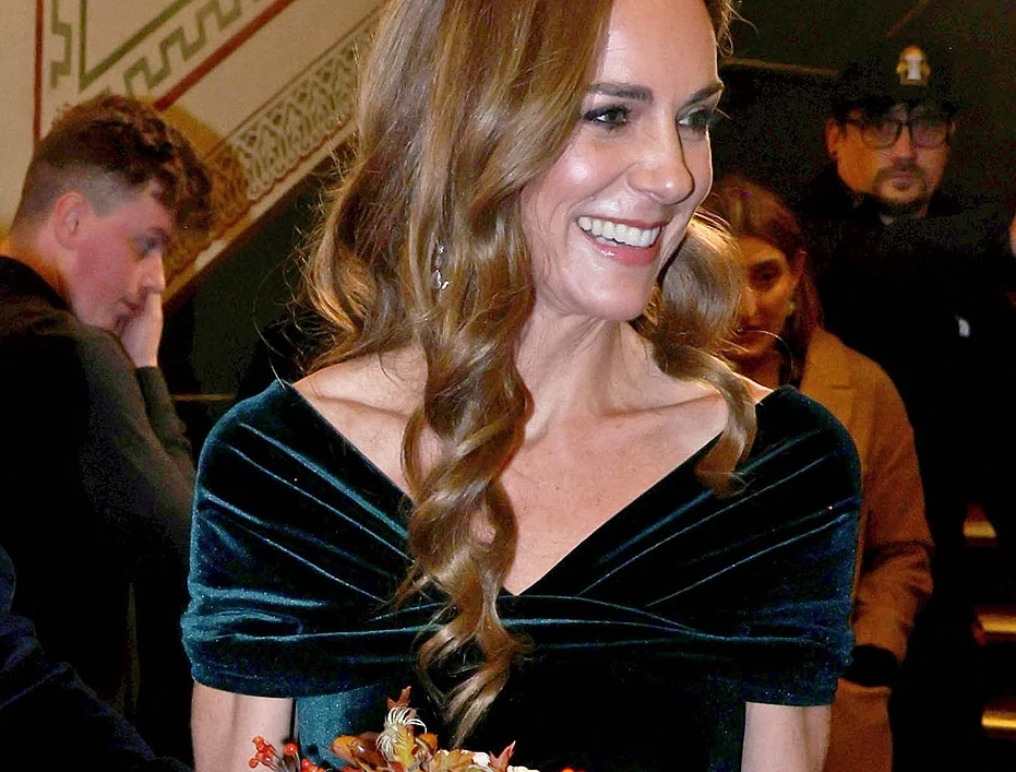 Kobieta w eleganckiej, ciemnozielonej sukni z bukietem kwiatów w dłoniach, uśmiechnięta, w tle widoczni są inni ludzie i elementy wystroju wnętrza, w tym ozdobna ściana.