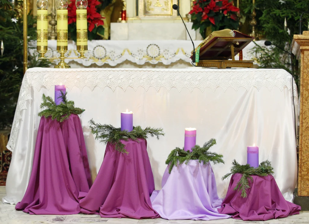Cztery świece adwentowe w kolorach fioletu i różu ustawione przed ołtarzem, oplecione gałązkami iglastymi i otoczone materiałem, w tle ołtarz i księga liturgiczna.