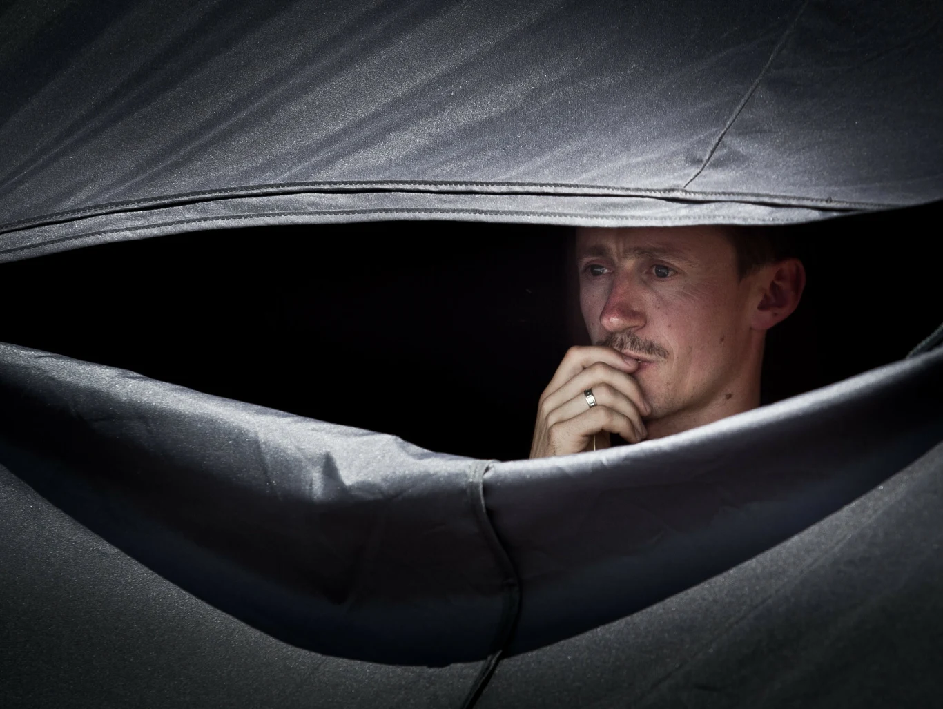 Adam Małysz w 2011 roku przed jednym z rajdów Mężczyzna patrzący przez rozcięcie w ciemnym materiale, głęboko zamyślony, opiera dłoń na brodzie, światło wydobywa z mroku jedynie jego twarz i rękę.