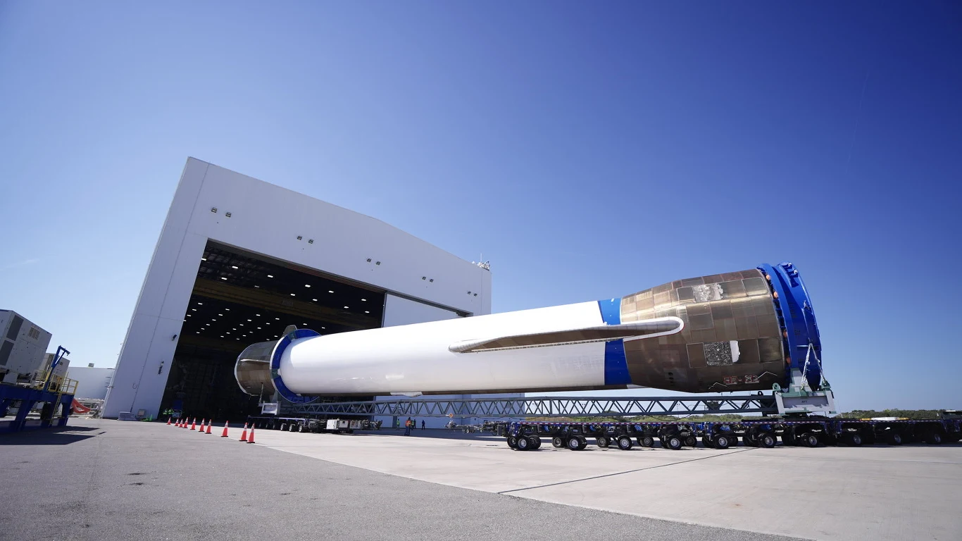 New Glenn jak Starship? Nowa rakieta Blue Origin będzie większa. Duża rakieta transportowana na platformie przed hangarem, jasnoniebieskie niebo, nowoczesna infrastruktura przemysłowa i elementy techniczne wokół miejsca załadunku.