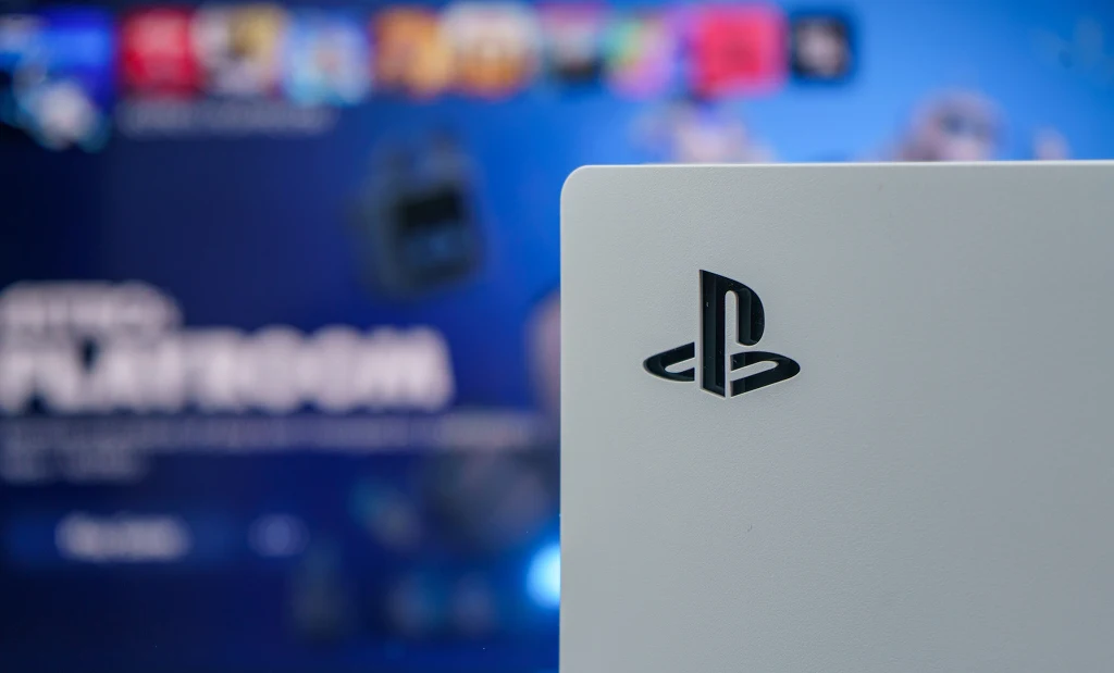 Biała konsola PlayStation z charakterystycznym czarnym logo na przednim panelu na tle rozmytego ekranu telewizora z kolorowym interfejsem użytkownika konsoli.