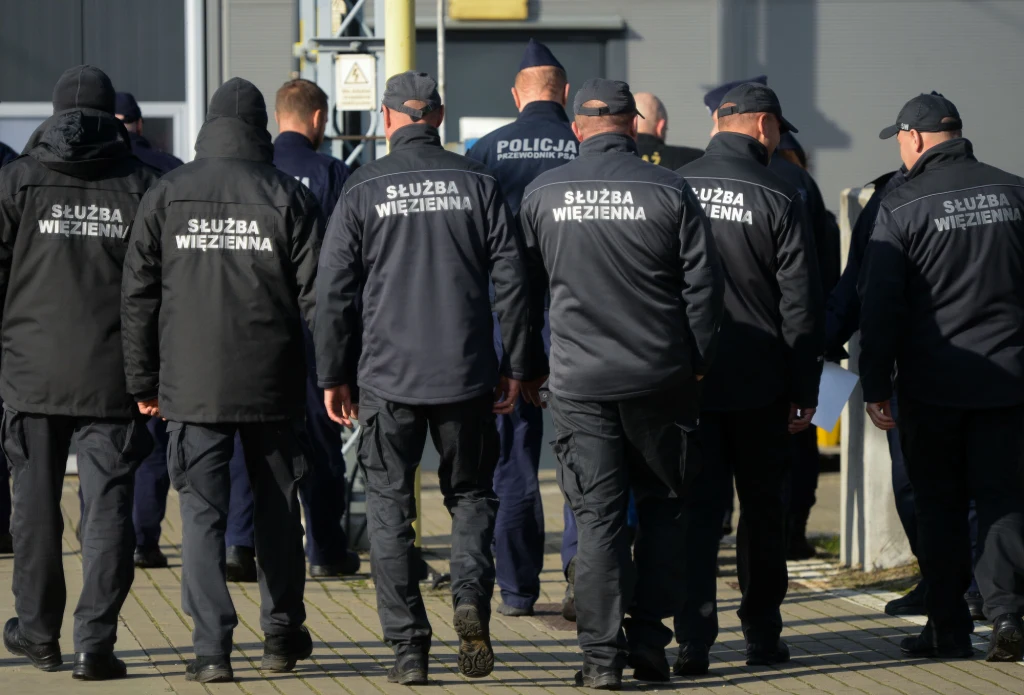 Służba Więzienna otrzyma dodatek mieszkaniowy Grupa funkcjonariuszy służby więziennej w ciemnych mundurach idzie razem po chodniku w kierunku budynku, ich plecy są widoczne, a na kurtkach widnieje napis 'Służba Więzienna'.