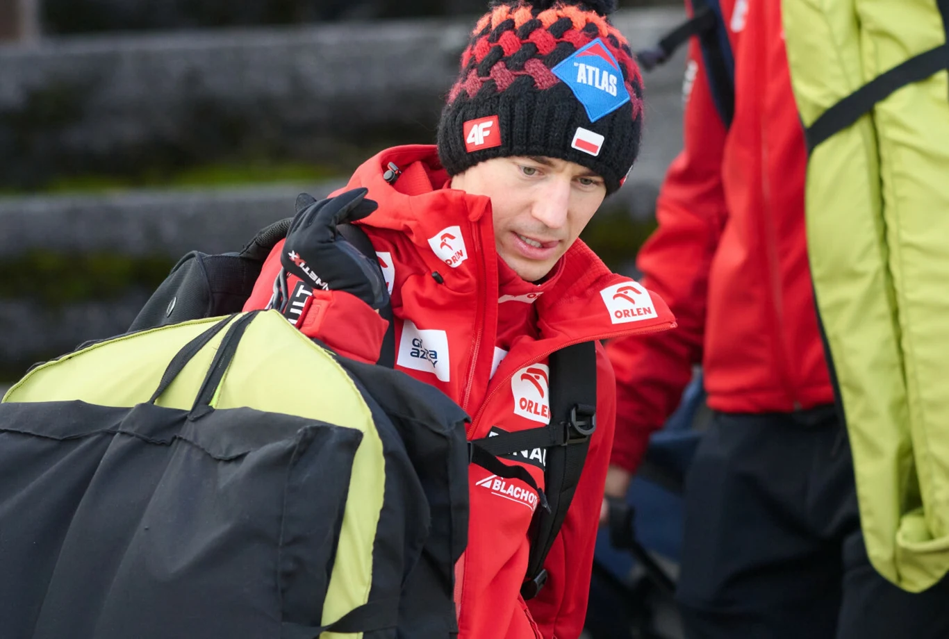 Kamil Stoch Mężczyzna w czerwonym stroju sportowym z licznymi logotypami, w tym Orlen i Atlas, ma na głowie czapkę, niesie dużą torbę sportową na ramieniu, w tle widać osobę w zielonej kurtce.