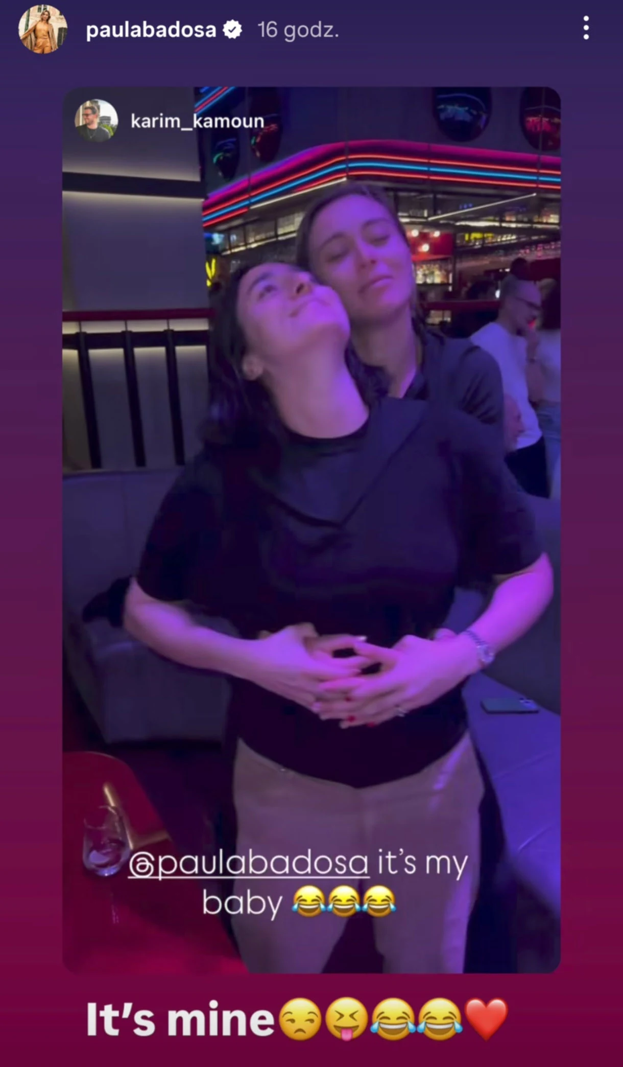 Dwie osoby obejmują się ciepło w kolorowo oświetlonym wnętrzu, tworząc serdeczną i radosną atmosferę. W tle widoczny lokal z jasnymi światłami oraz inni ludzie, co sugeruje miejsce publiczne o luźnej atmosferze.