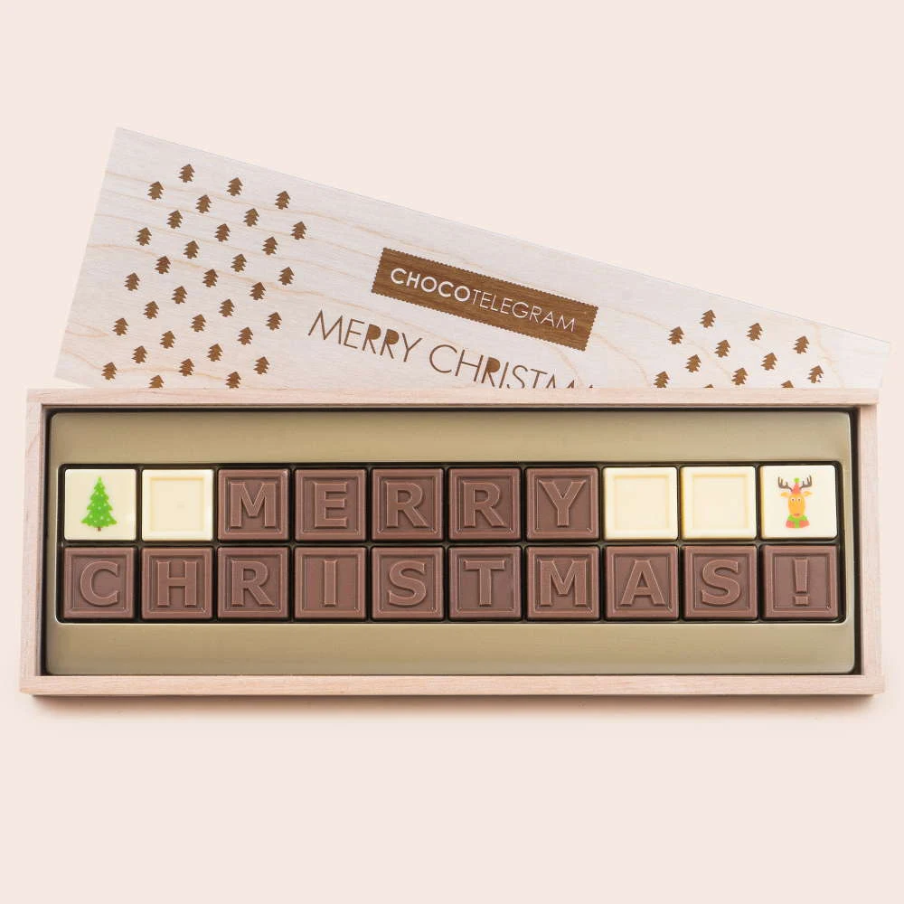Czekoladki ułożone w pudełku w kształcie prostokątnym, tworzące napis Merry Christmas, ozdobione rysunkiem choinki oraz renifera na końcach, elegancko zapakowane w drewnianym pudełku z napisem CHOCOTELEGRAM.