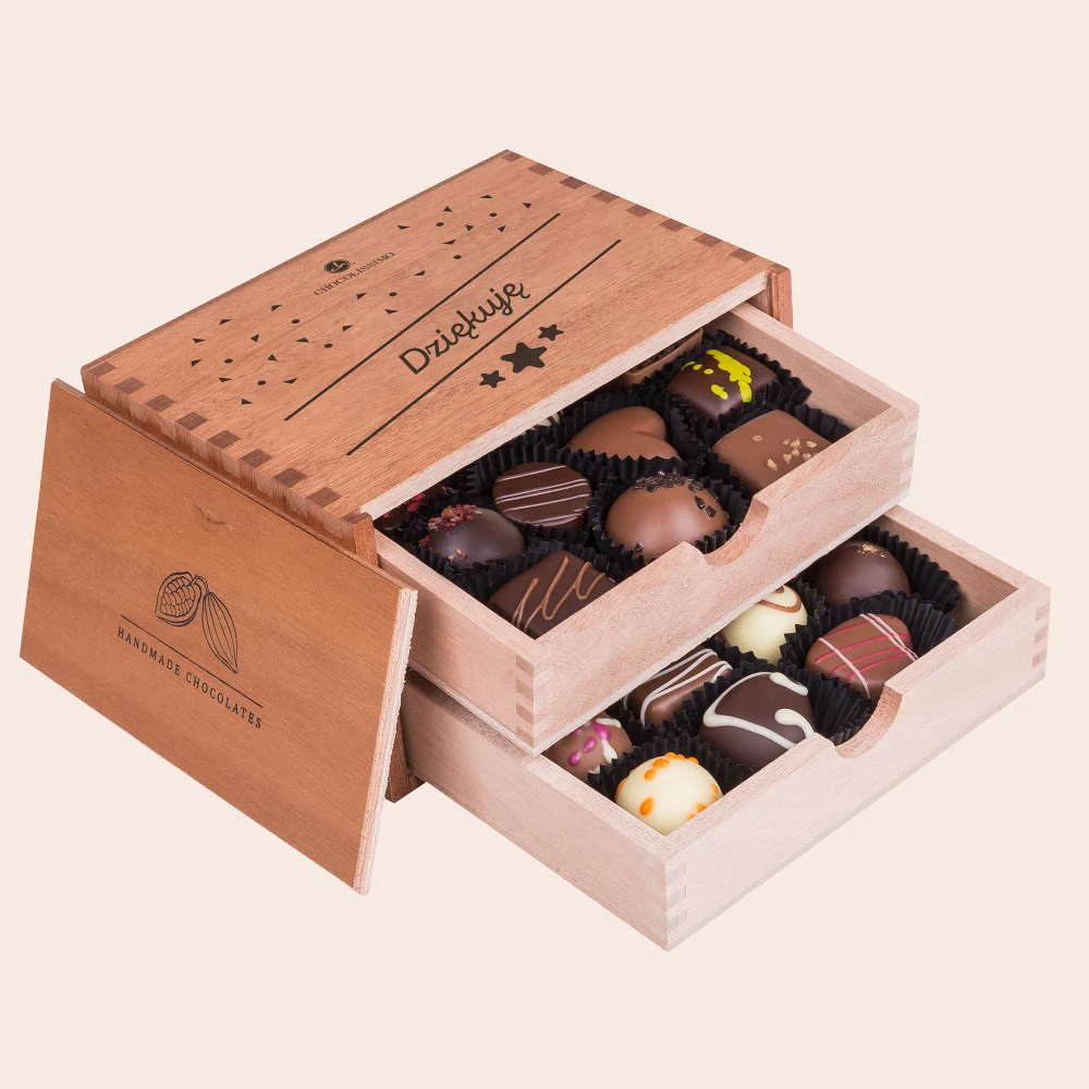 Otwarta, elegancka, drewniana szkatułka z napisem 'Dziękuję', w której znajdują się dwie szuflady wypełnione różnorodnymi, ozdobionymi pralinkami czekoladowymi.