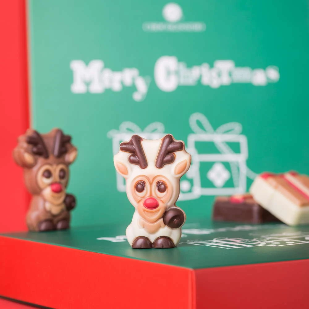 Czekoladowa figurka renifera z dużymi oczami i czerwonym noskiem ustawiona na kolorowym pudełku z napisem Merry Christmas oraz świątecznym nadrukiem prezentowym, w tle druga podobna figurka i kawałki czekolady.